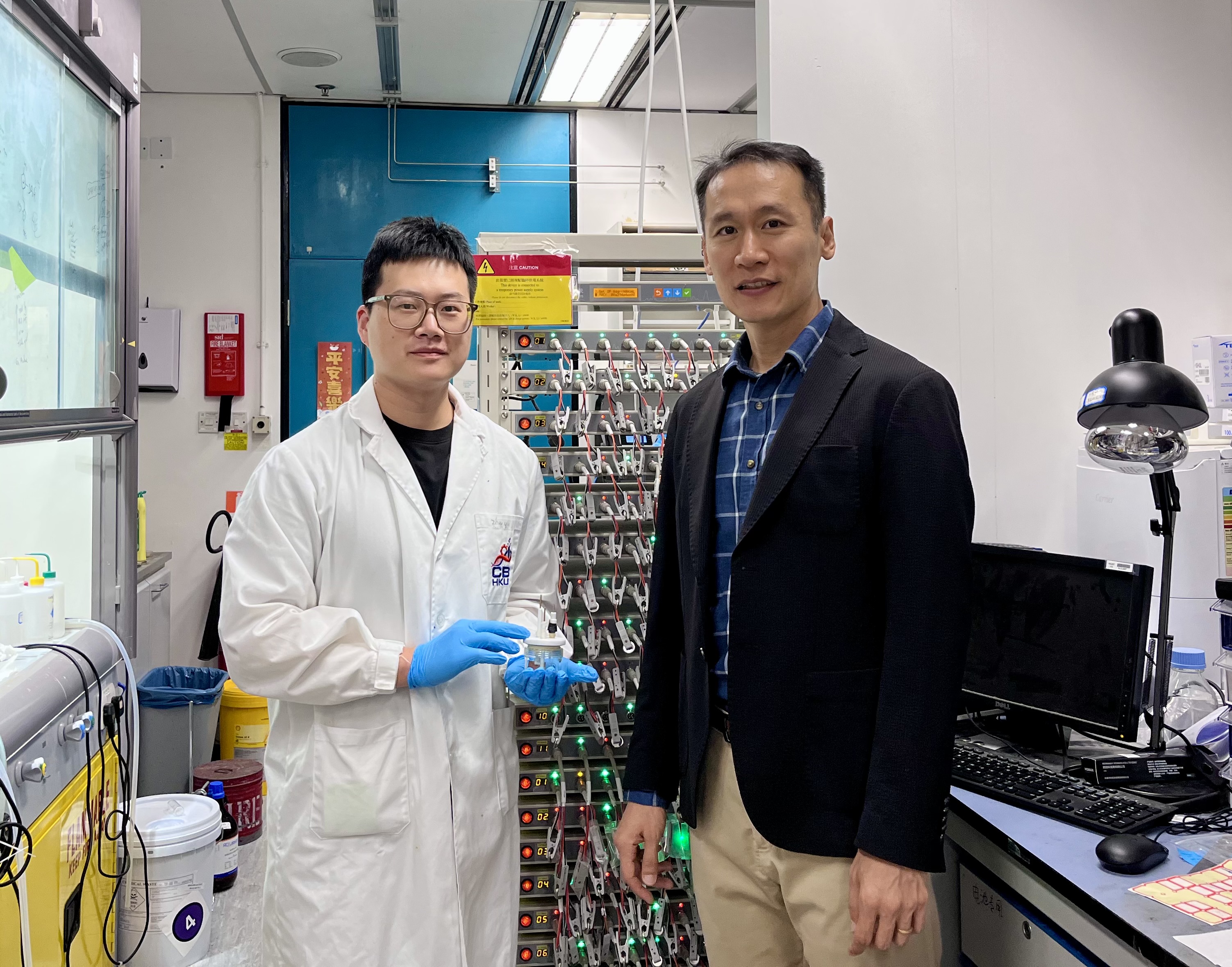 科大化學及生物工程學系副教授兼論文的通訊作者金允燮教授（右）及其博士生兼論文第一作者尹卓毓（左；手持電化學電池模具）站在電池充放電循環儀旁合影。